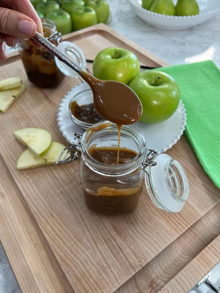 A spoon drizzling glossy vegan caramel sauce into a glass jar next to green apples