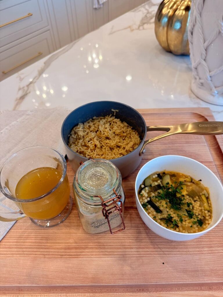Vegan chicken soup base jar next to a mug of broth, seasoned rice and a bowl of soup on a wooden board