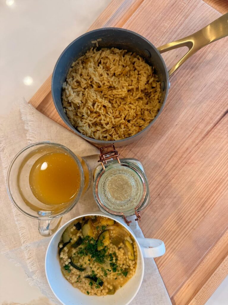 Homemade vegan chicken soup base in a glass jar with a mug of broth, a pot of seasoned rice and a bowl of pastina soup