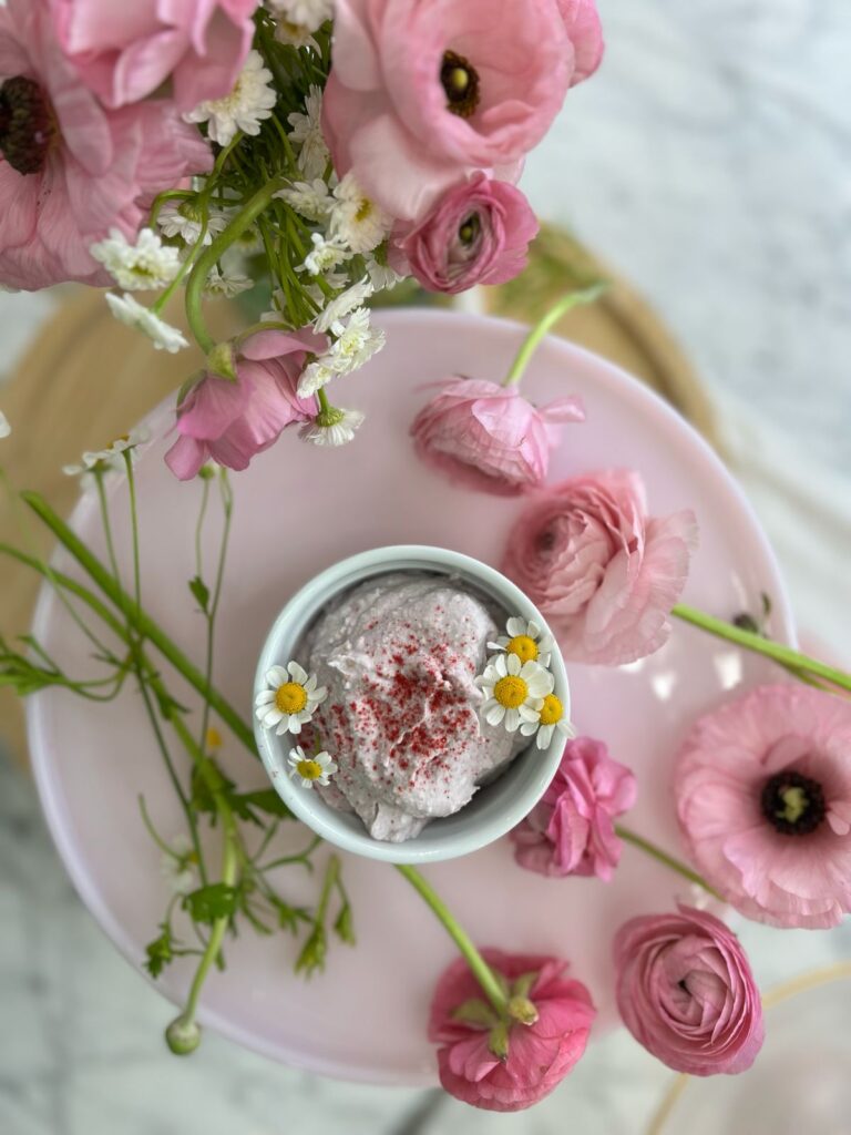 A ramekin of mousse topped with daisies on a pink cake stand surrounded by pink ranunculus flowers